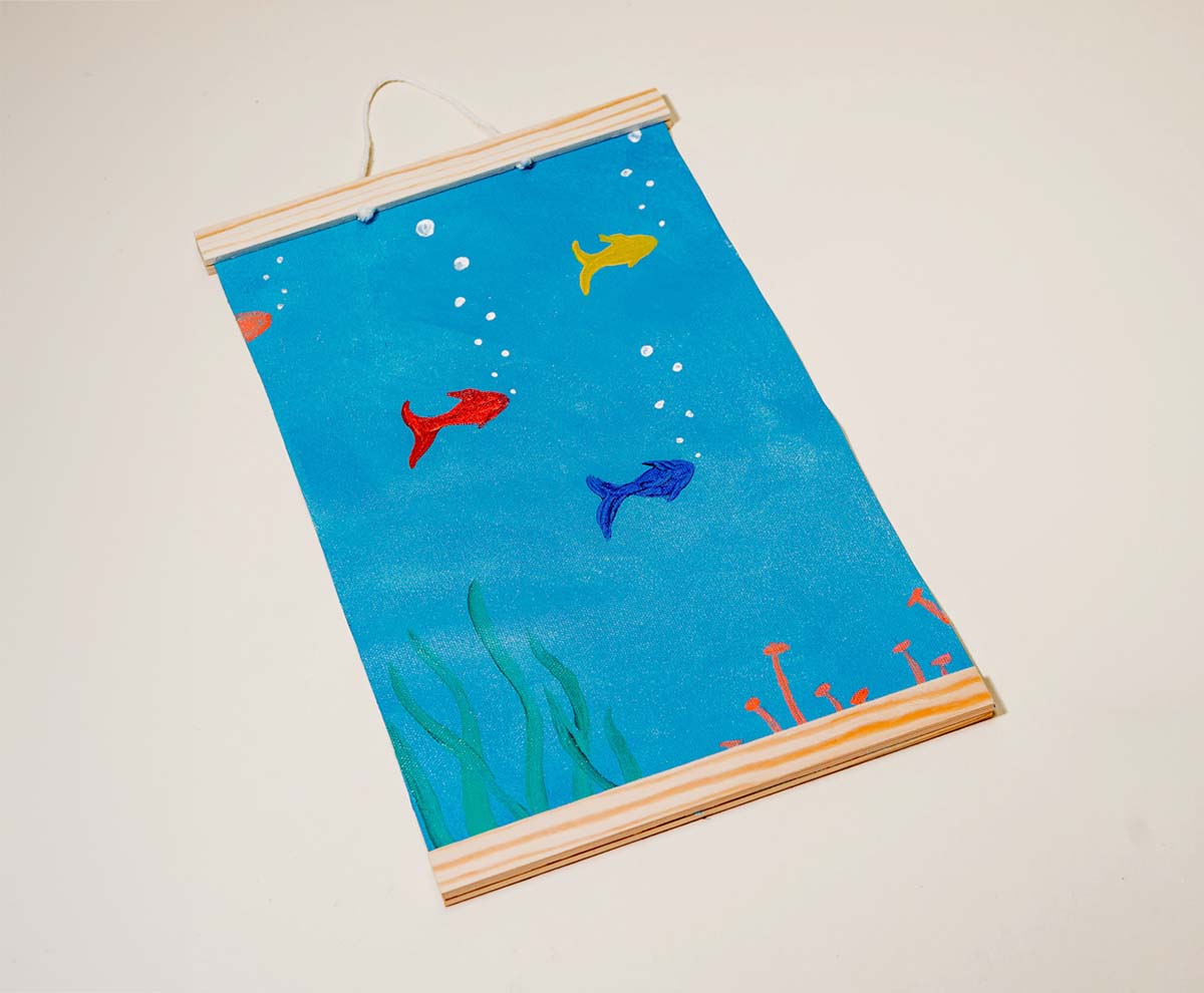 A handmade wall hanging with a vivid blue background featuring three small fish in yellow, red and blue with sea plants framed with wooden rods made with Sculpd Kids Canvas Painting Kit