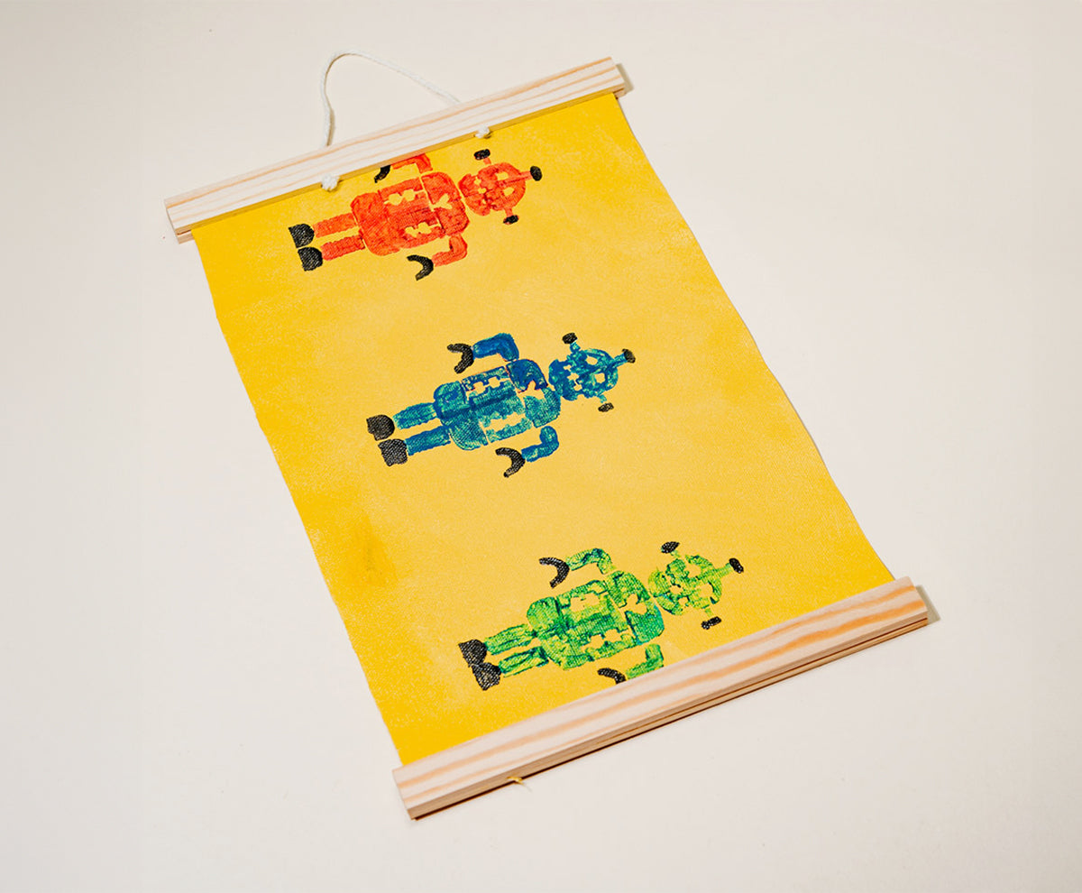 A handmade wall hanging with a yellow background featuring three stamped robots in red, blue, and green, framed with wooden rods made with Sculpd Kids Canvas Painting Kit