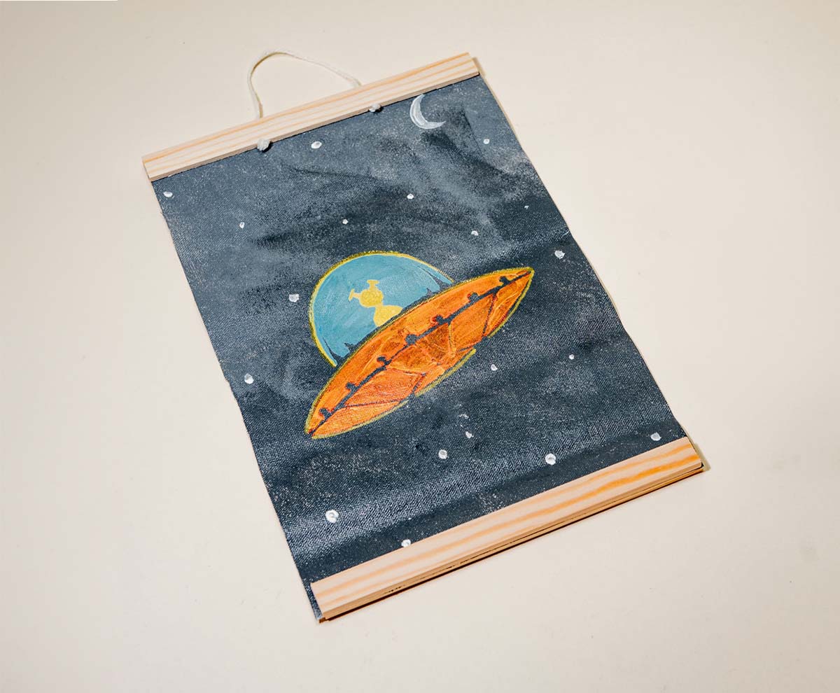 A handmade wall hanging with a dark blue background featuring a moon and a spaceship framed with wooden rods made with Sculpd Kids Canvas Painting Kit