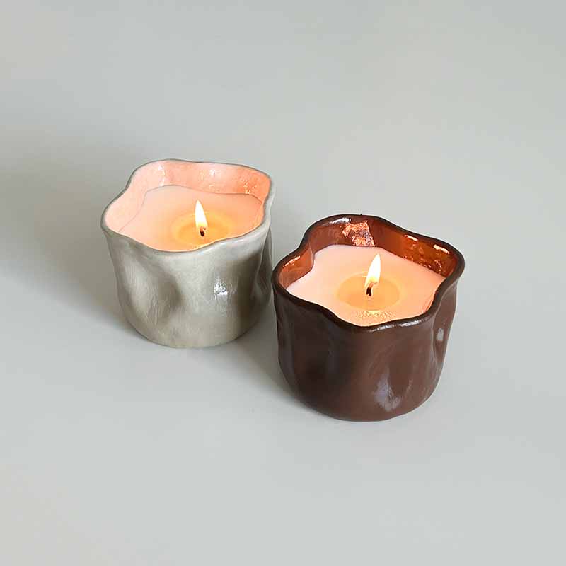 Two handmade candles burn inside organic-shaped clay vessels, one in earthy brown and the other in soft beige, made with Sculpd Candle Making Kit