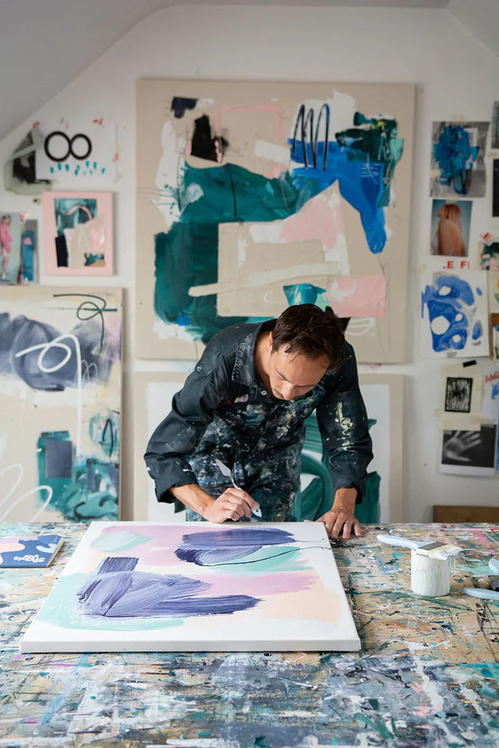 Man wearing paint-splattered overalls leaned over and painting on a canvas on a table