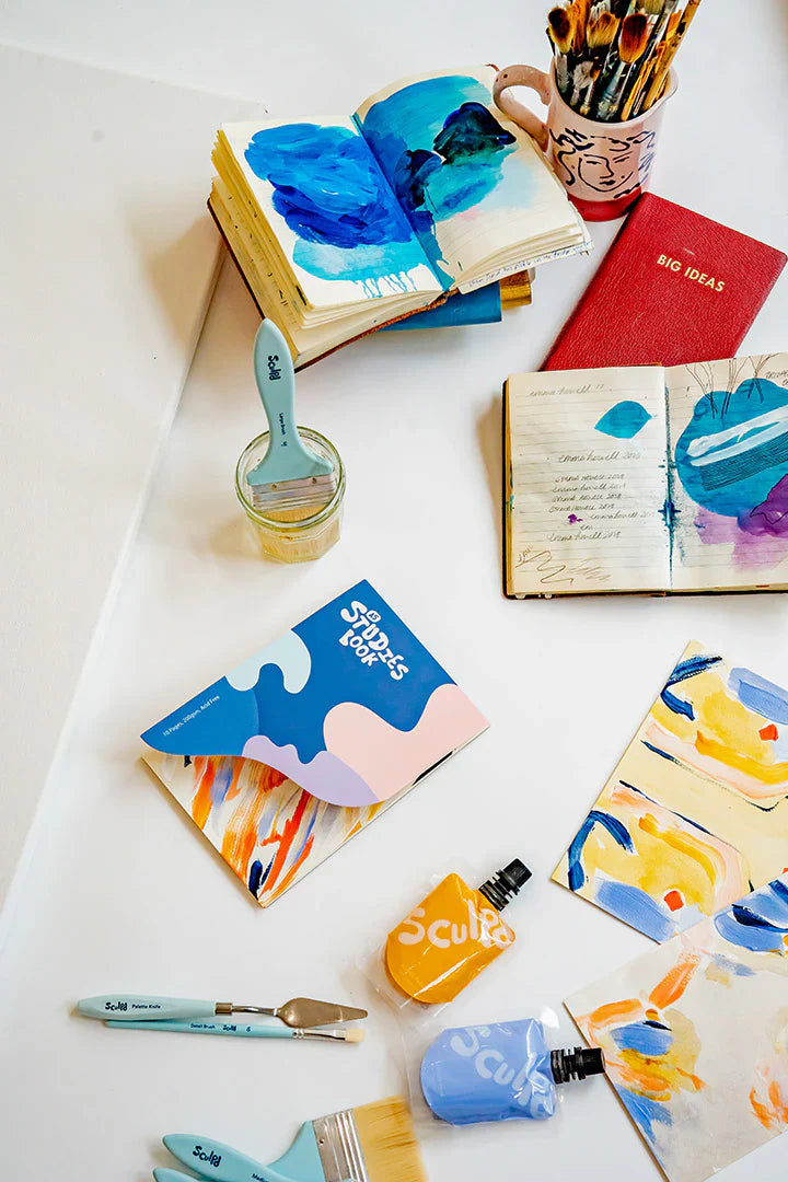 An artistic workspace with open sketchbooks, paintbrushes, and Sculpd-branded paint pouches, featuring vibrant blue and yellow abstract paintings